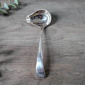 May include: A silver gravy ladle with a curved handle and a rounded bowl. The ladle is made of a shiny, reflective metal and is set against a wooden background. The ladle is designed for serving sauces and gravies.