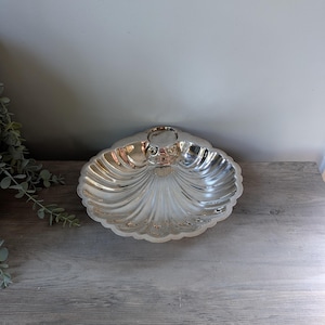 May include: A silver-toned metal dish shaped like a scallop shell. The dish features a reflective, polished surface with deep, radiating fluted grooves that extend from a central circular hinge point to the scalloped, wavy outer edge.