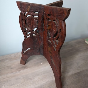 May include: A dark brown, wooden decorative stand with carved floral designs. The stand has four legs and a folding design. The wood has a distressed finish, adding to its rustic charm. The stand is likely used for displaying small items.