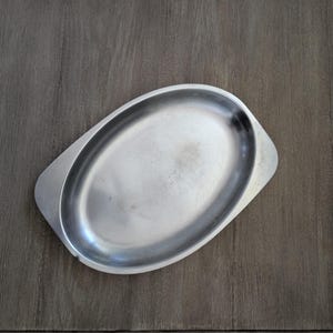 May include: A silver-colored, oval-shaped serving tray with a slightly raised rim. The tray has a handle on each end and is made of a shiny metal. The tray is empty and sits on a dark wooden surface.