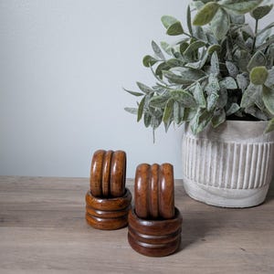 May include: Two sets of wooden napkin rings. Each set includes four rings stacked on a matching base. The rings and bases are a warm, medium brown color. A potted plant with green leaves is in the background.