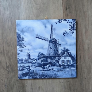 May include: Blue and white ceramic tile featuring a rural landscape with windmills, houses, and a horse. The artwork depicts a traditional Dutch scene, with a windmill as the focal point. The tile is square and has a matte finish.
