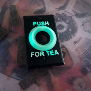 May include: A black plastic button with a glowing green circle and the text "PUSH FOR TEA" printed on it.