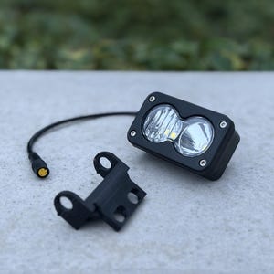 May include: A rectangular black LED auxiliary light with two circular lenses and four silver mounting screws, accompanied by a separate black plastic mounting bracket and a short black electrical cable with a circular connector plug.