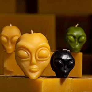 May include: Four alien-shaped candles, two yellow, one green, and one black, are arranged on a yellow surface. The candles are all in the shape of an alien head with large eyes and a small mouth.