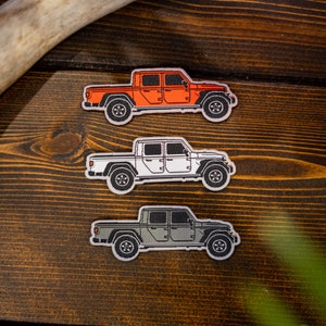 May include: Three embroidered patches of a Jeep Gladiator pickup truck in orange, white, and grey on a wooden surface.