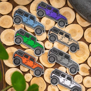 以下が含まれることがあります： 青、紫、緑、ベージュ、グレー、オレンジ、白など、さまざまな色のカラフルなジープパッチのコレクション。各パッチには、ジープ車の詳細な刺繍デザインが施されています。パッチは木製の表面に配置されています。