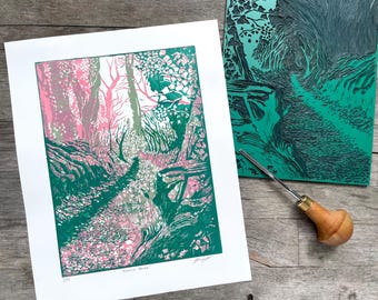 May include: A green and pink linocut print of a path through a forest. The print is signed and numbered. The image shows the linocut block next to the print.