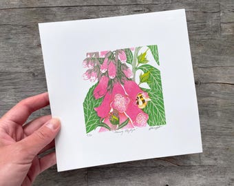May include: A square print depicting a pink foxglove flower with green foliage and a bee. The artwork is titled "Nancy Blythyn" and is numbered 1/50. The print is held against a wooden surface.