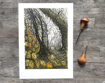 May include: A black and yellow linocut print of a forest scene with a path. The print is on white paper, with two wood-handled carving tools resting on a wooden surface.