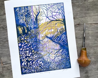 May include: A blue and yellow linocut print depicting a river scene. The river is in the centre of the image and is surrounded by trees and foliage. The print is titled "River Dreaming".