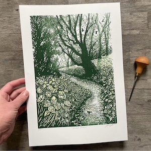 May include: A green and white linocut print of a woodland stream with a single tree in the background. The print is titled "Woodland Stream" and signed by the artist.