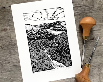 May include: Monochrome landscape print showing a valley with a river, mountains, and clouds. Two wood carving tools with brass details are beside the print. The print is on white paper.