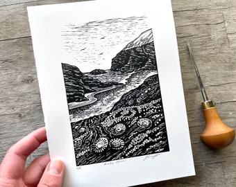 May include: Black and white linocut print depicting a rocky shore with a winding river leading to a mountain range in the distance. The print is titled "Rocky Shore" and signed by the artist.