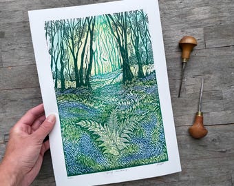 May include: A linocut print titled Spring Sanctuary featuring a forest scene with tall dark green trees, a foreground of bluebell flowers, and a central fern. The artwork uses shades of green, blue, and yellow, with two wooden carving tools nearby.