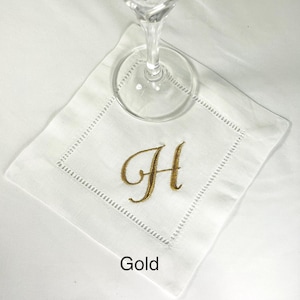 K&ouml;nnte beinhalten: Wei&szlig;es Cocktail-Servietten aus Leinen mit einem goldgestickten Buchstaben "H". Die Serviette hat eine dekorative, gen&auml;hte Bord&uuml;re. Ein klares Weinglas liegt auf der Serviette. Das Wort "Gold" ist unten aufgedruckt.