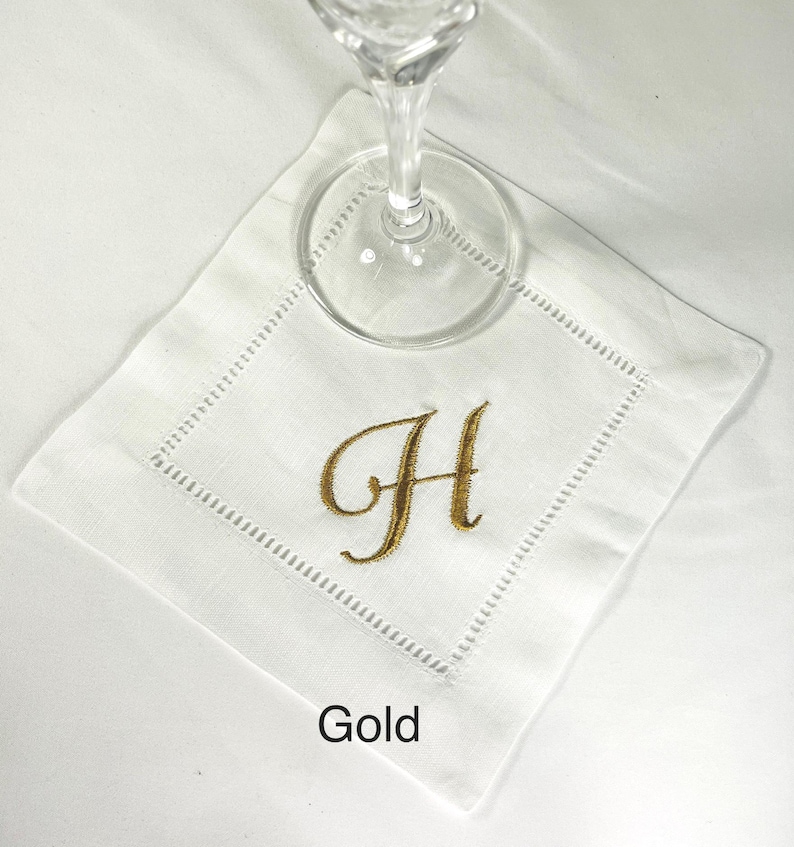 K&ouml;nnte beinhalten: Wei&szlig;es Cocktail-Servietten aus Leinen mit einem goldgestickten Buchstaben "H". Die Serviette hat eine dekorative, gen&auml;hte Bord&uuml;re. Ein klares Weinglas liegt auf der Serviette. Das Wort "Gold" ist unten aufgedruckt.