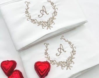 May include: Two folded white pillowcases featuring the text Old Lace and an embroidered beige floral wreath surrounding the letter A. Three red heart-shaped candies wrapped in foil are placed near the bottom pillowcase.