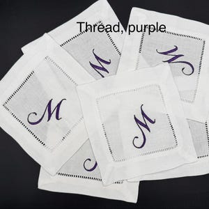 K&ouml;nnte beinhalten: Eine Sammlung wei&szlig;er Leinenuntersetzer, jeder mit einem lila Buchstaben "M" bestickt. Die Untersetzer haben eine quadratische Form mit einem dekorativen, gen&auml;hten Rand. Der Text "Thread, purple" ist im Bild zu sehen.