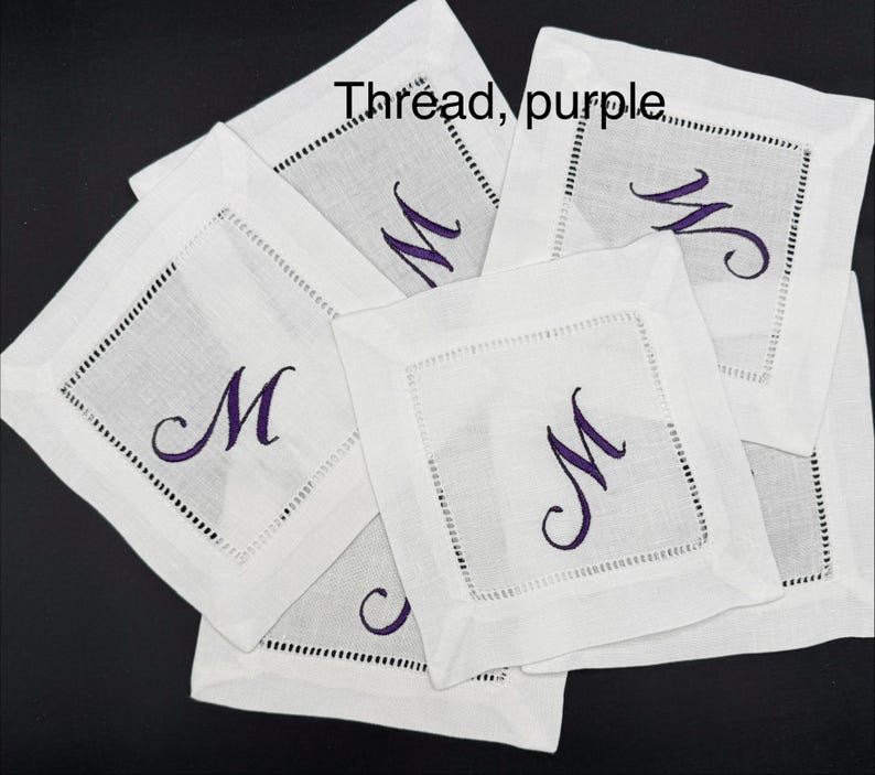 K&ouml;nnte beinhalten: Eine Sammlung wei&szlig;er Leinenuntersetzer, jeder mit einem lila Buchstaben "M" bestickt. Die Untersetzer haben eine quadratische Form mit einem dekorativen, gen&auml;hten Rand. Der Text "Thread, purple" ist im Bild zu sehen.