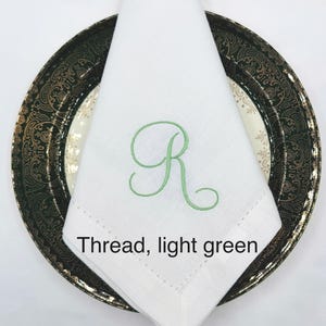 May include: A white linen napkin with a light green embroidered letter "R" is folded on a decorative plate. The plate has a black and gold rim. The text "Thread, light green" is below the napkin.