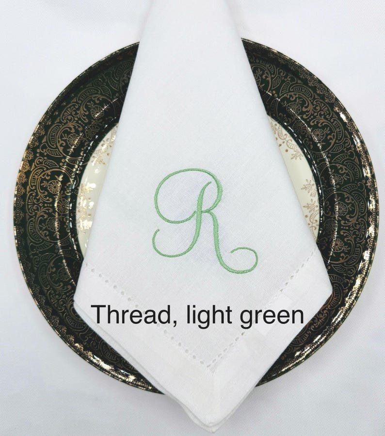 May include: A white linen napkin with a light green embroidered letter "R" is folded on a decorative plate. The plate has a black and gold rim. The text "Thread, light green" is below the napkin.