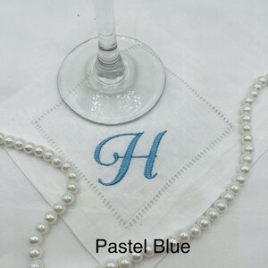 K&ouml;nnte beinhalten: Wei&szlig;es besticktes Taschentuch mit einem pastellblauen Buchstaben "H" und einer Perlenkette. Das Taschentuch hat eine dekorative, gen&auml;hte Bord&uuml;re. Die Worte "Pastel Blue" sind am unteren Rand des Bildes aufgedruckt. Ein Weinglas ist im Hintergrund.