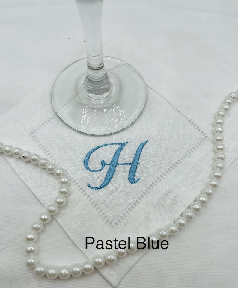 K&ouml;nnte beinhalten: Wei&szlig;es besticktes Taschentuch mit einem pastellblauen Buchstaben "H" und einer Perlenkette. Das Taschentuch hat eine dekorative, gen&auml;hte Bord&uuml;re. Die Worte "Pastel Blue" sind am unteren Rand des Bildes aufgedruckt. Ein Weinglas ist im Hintergrund.