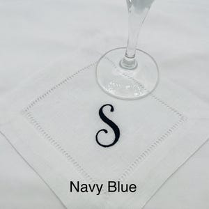 K&ouml;nnte beinhalten: Wei&szlig;es Cocktail-Servietten aus Leinen mit einem marineblauen, gestickten Buchstaben "S". Die Serviette hat eine dekorative, gen&auml;hte Bord&uuml;re und die Worte "Navy Blue" sind darunter gedruckt. Ein klares Glas liegt darauf.