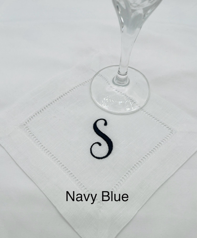 K&ouml;nnte beinhalten: Wei&szlig;es Cocktail-Servietten aus Leinen mit einem marineblauen, gestickten Buchstaben "S". Die Serviette hat eine dekorative, gen&auml;hte Bord&uuml;re und die Worte "Navy Blue" sind darunter gedruckt. Ein klares Glas liegt darauf.