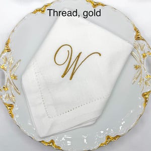 May include: A white linen napkin with a gold embroidered letter "W" is displayed on a white plate with a gold rim. The text "Thread, gold" is visible in the image. The napkin is folded and has a decorative stitched border.