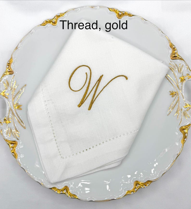 May include: A white linen napkin with a gold embroidered letter "W" is displayed on a white plate with a gold rim. The text "Thread, gold" is visible in the image. The napkin is folded and has a decorative stitched border.