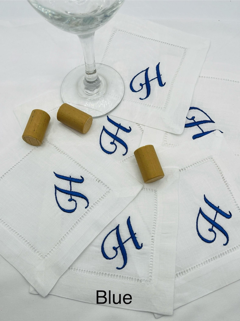 K&ouml;nnte beinhalten: Quadratische wei&szlig;e Leinenuntersetzer mit einem blauen Buchstaben "H" bestickt. Die Untersetzer sind mit Weinkorken und einem Weinglas angeordnet. Das Wort "Blue" befindet sich am unteren Rand des Bildes.