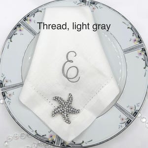 May include: A white linen napkin with a light gray embroidered letter "E" and a decorative rhinestone starfish on a floral patterned plate. The text "Thread, light gray" is above the napkin.