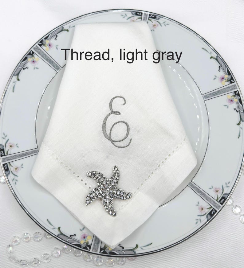 May include: A white linen napkin with a light gray embroidered letter "E" and a decorative rhinestone starfish on a floral patterned plate. The text "Thread, light gray" is above the napkin.