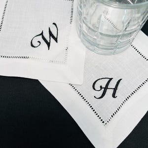 K&ouml;nnte beinhalten: Wei&szlig;e Cocktailservietten aus Leinen mit schwarz gestickten Initialen "W" und "H". Jede Serviette hat eine dekorative, gen&auml;hte Bord&uuml;re. Ein klares Glas steht auf einer der Servietten.