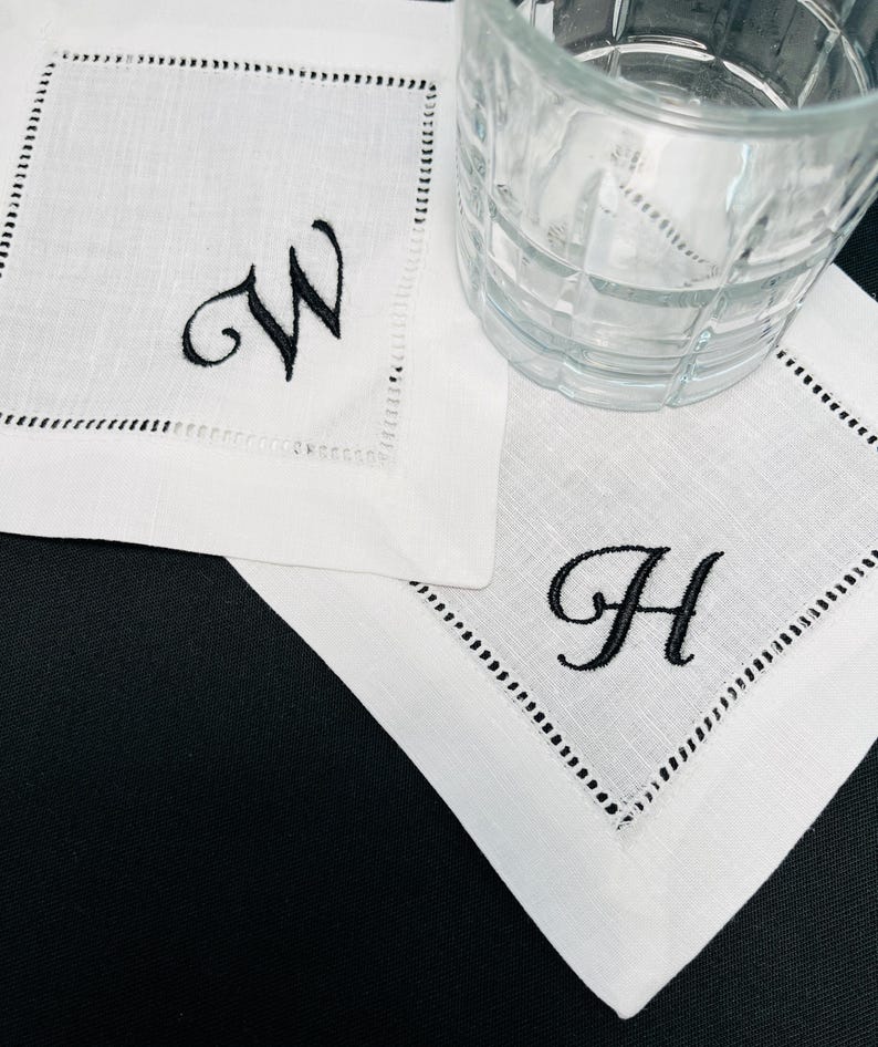 K&ouml;nnte beinhalten: Wei&szlig;e Cocktailservietten aus Leinen mit schwarz gestickten Initialen "W" und "H". Jede Serviette hat eine dekorative, gen&auml;hte Bord&uuml;re. Ein klares Glas steht auf einer der Servietten.