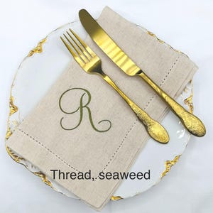May include: A gold-colored fork and knife set on a white plate with gold trim, next to a beige linen napkin embroidered with a green letter "R". The napkin has the text "Thread, seaweed".