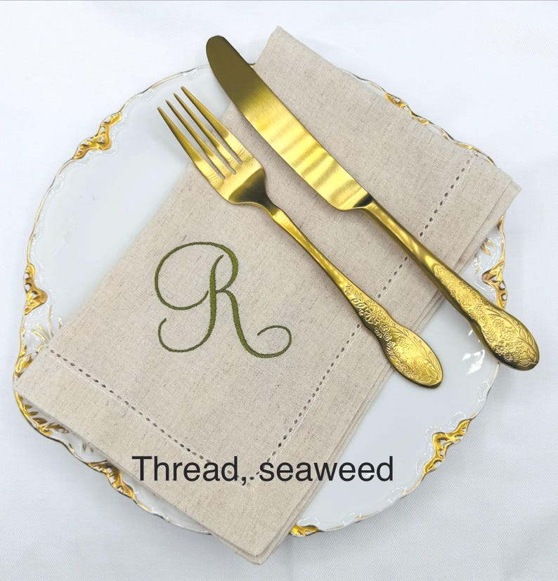 May include: A gold-colored fork and knife set on a white plate with gold trim, next to a beige linen napkin embroidered with a green letter "R". The napkin has the text "Thread, seaweed".