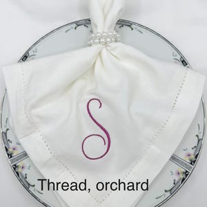 May include: A white linen napkin with a stitched border and a magenta embroidered "S" sits on a floral-patterned plate. A pearl beaded napkin ring holds the napkin. The text "Thread, orchard" is at the bottom.