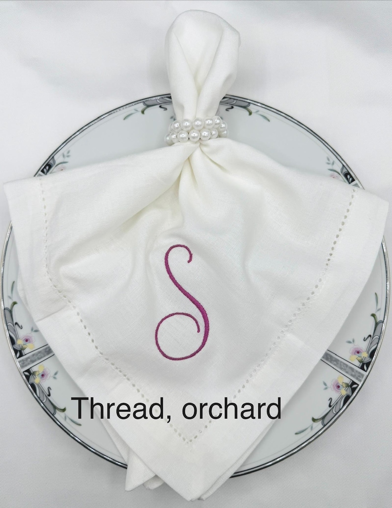 May include: A white linen napkin with a stitched border and a magenta embroidered "S" sits on a floral-patterned plate. A pearl beaded napkin ring holds the napkin. The text "Thread, orchard" is at the bottom.