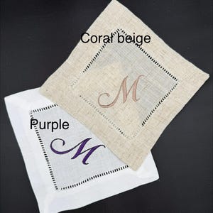 K&ouml;nnte beinhalten: Zwei quadratische Leinenuntersetzer, einer in Korallenbeige und einer in Wei&szlig;. Jeder Untersetzer hat einen gestickten Buchstaben "M" in einer Komplement&auml;rfarbe. Der korallenbeige Untersetzer hat ein braunes "M", und der wei&szlig;e Untersetzer hat ein lila "M".