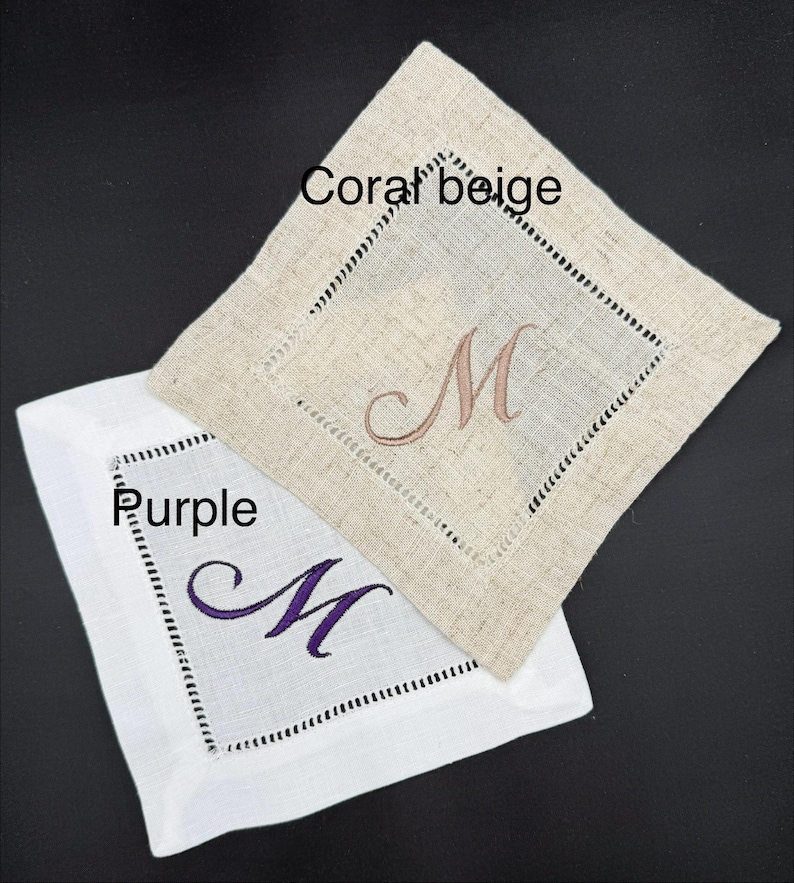 K&ouml;nnte beinhalten: Zwei quadratische Leinenuntersetzer, einer in Korallenbeige und einer in Wei&szlig;. Jeder Untersetzer hat einen gestickten Buchstaben "M" in einer Komplement&auml;rfarbe. Der korallenbeige Untersetzer hat ein braunes "M", und der wei&szlig;e Untersetzer hat ein lila "M".