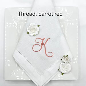 May include: A white linen napkin with a carrot red embroidered letter "K" and a decorative stitched border. The napkin is folded on a white square plate and adorned with white paper roses. The text "Thread, carrot red" is visible.
