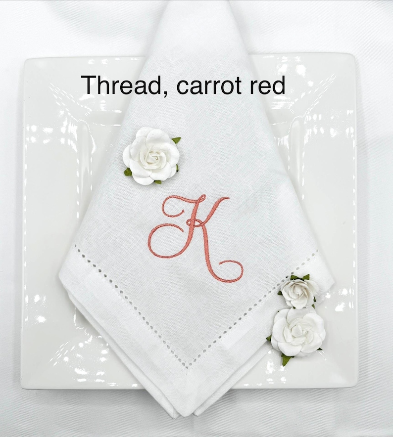 May include: A white linen napkin with a carrot red embroidered letter "K" and a decorative stitched border. The napkin is folded on a white square plate and adorned with white paper roses. The text "Thread, carrot red" is visible.