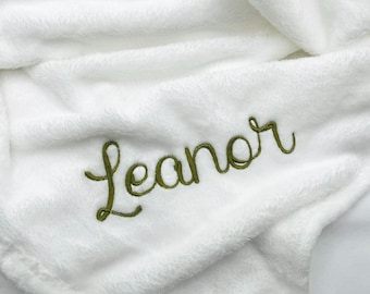 May include: A soft white plush blanket featuring the embroidered name Leanor in seaweed green thread. The text Thread is seaweed is printed in black above the name, indicating the color of the embroidery thread used for the personalization.