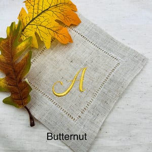 K&ouml;nnte beinhalten: Eine beige Leinen-Serviette mit einem goldgestickten Buchstaben "A" und einem dekorativen, gen&auml;hten Rand. Das Wort "Butternut" ist unten aufgedruckt. Herbstbl&auml;tter in Gelb- und Braunt&ouml;nen liegen neben der Serviette.