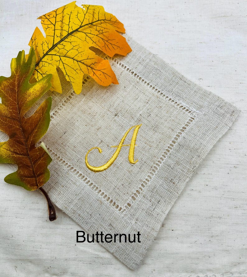 K&ouml;nnte beinhalten: Eine beige Leinen-Serviette mit einem goldgestickten Buchstaben "A" und einem dekorativen, gen&auml;hten Rand. Das Wort "Butternut" ist unten aufgedruckt. Herbstbl&auml;tter in Gelb- und Braunt&ouml;nen liegen neben der Serviette.