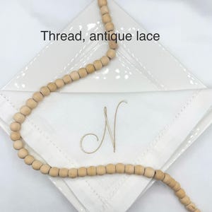 May include: A white linen napkin with an embroidered "N" is displayed on a white plate. A strand of wooden beads is draped across the napkin. The text "Thread, antique lace" is above the napkin.