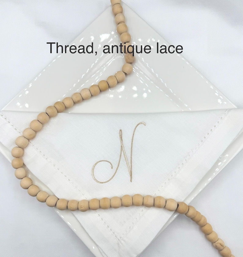 May include: A white linen napkin with an embroidered "N" is displayed on a white plate. A strand of wooden beads is draped across the napkin. The text "Thread, antique lace" is above the napkin.